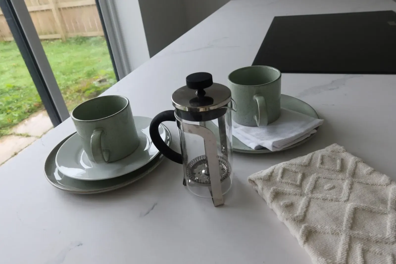 A French press and two coffee mugs on a bright kitchen counter in a warm, comfortable home